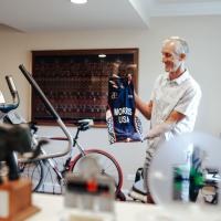 Doug, wearing a white button-down, smiles as he holds up a cycling jersey that says MORRIS USA. He is inside his home, in a space with a bike and some workout equipment.