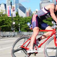 An action shot of Doug during a triathlon. He is wearing an American-flag-print unitard and is riding a red bicycle around a curve.