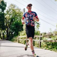 Doug, wearing the flag-print jersey, black bike shorts, and red and white striped socks, grins as he runs down a sidewalk. He is wearing a baseball hat and sunglasses.