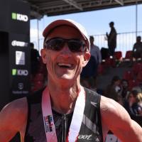 Doug smiles widely after receiving a medal at a triathlon. He is wearing a black unitard, sunglasses, and a baseball hat.