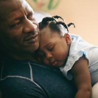 A photo of Betty sleeping on her grandfather’s chest. He is an older Black man with short graying dark hair. He is wearing a blue long-sleeve shirt with white stitching.