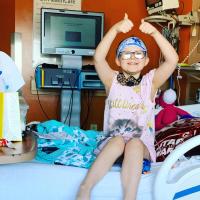 A photo of Ellie in her hospital room where she is sitting upright on the bed with both arms in the air giving two thumbs ups. Her room is full of colorful blankets, stuffed animals, and a yellow gift bag on her bed.