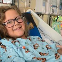 A close-up photo of Ellie laying in her hospital bed smiling at the camera while giving the Star Trek “live long and prosper” hand symbol for the photo. She is wearing a blue gown with cartoon tigers on it.