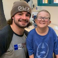 Ellie and Kash Daniel, a young man with an athletic build, brown hair, and a beard wearing a brown beanie and a gray and black t-shirt, sit together and smile for a picture.