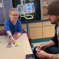 Ellie and Kash sit together at a table in the hospital and play Uno together.