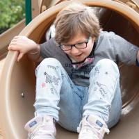 A photo of Ellie at the bottom of a tunnel slide with her hands grabbing the outside of the slide while she enthusiastically smiles. She is wearing a gray t-shirt, jeans, and light pink sneakers.