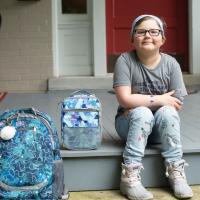 A photo of Ellie sitting on her front porch with her hands on her legs next to her school backpack and lunchbox.