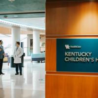 A photo showing the front lobby of Kentucky Children’s Hospital, where two doctors are conversing. A plaque reading ‘Kentucky Children’s Hospital’ with the UK HealthCare logo is in the forefront of the photo.