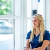 Emma, wearing dark blue scrubs, sits at a chair and looks out a window. Her blonde hair is loose around her shoulders.