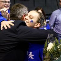 Emma, with a small blue Wildcat head painted on her cheek, hugs a man wearing a suit. She is holding a bouquet of flowers. People in the background are applauding.