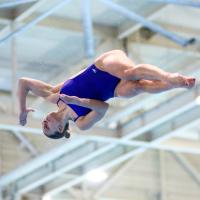 Emma is mid-dive, wearing a blue one-piece swimsuit. She is midair, turning sideways and twisting with her legs straight out and her toes pointed.