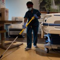 A person dressed in scrubs, gloves and a face mask is cleaning a patient room using a dust mop.