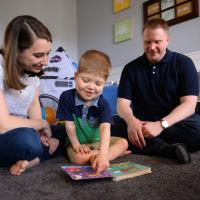 A photo of Everett and his parents Claire and Daniel sitting on the floor and reading a children’s book together. Claire is a white woman with medium length dark brown hair. She is wearing a white short-sleeve shirt with a geometric pattern on it and a pair of blue jeans. Daniel is a white man with short red hair. He is wearing a short-sleeve navy polo and a pair of blue jeans.
