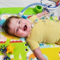 A photo of baby Everett smiling up at the camera as he lies on a mat. His trach tube can be seen connected to his neck.