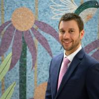 A photo of Dr. Kenneth Iverson smiling as he stands in front of a mosaic of a hummingbird and flowers. Dr. Iverson is a young white man with short brown hair. He is wearing a navy suit jacket with a white button-up shirt and a pink tie.