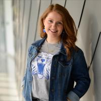 Fielden poses for a portrait in front of a wall at the UK student center. She is smiling and wearing a Wildcat tee, denim jacket, and blue and gold jewelry.