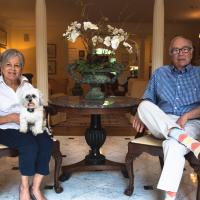 Gayle and her husband rest in chairs in their living room with their dog on Gayle's lap.