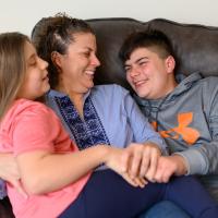 Geri sits on the couch embraced with her two children. Geri is wearing a blue striped shirt, her daughter is wearing a pink shirt, and her son is wearing a grey sweatshirt.