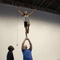 Gracie is lifted into the air by a young adult male practicing a cheerleading stunt. Her arms are raised and she is smiling.