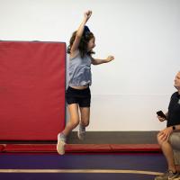 Gracie jumps enthusiastically on a gymnastics mat while her father, a middle aged man in a black polo shirt, watches her.