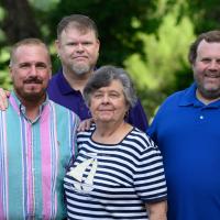 A photo of Helen standing in the middle of her three sons as they all pose for the camera. Her son Robert (left,) is an older white man with short brown hair and a brown beard. He is wearing a multicolor long-sleeve button-up. Her son Darren (middle,) is an older white man with short brown hair. He is wearing a short-sleeve navy polo, and multiple silver earrings. Her son Sean (right,) is an older white man with short curly brown hair. He is wearing a short-sleeve blue polo.