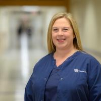 Allison Pinkston, a middle aged blonde white woman, stands smiling in the hospital hallway. She is wearing blue scrubs, as part of the required uniform.