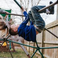 Henry and his younger sister, wearing a grey sweatshirt with blonde hair, are playing at a playground by climbing on a steel bar structure.