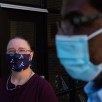 Marci Smith, a middle-aged white woman, is shown in focus on the left side of the photo. She has brown hair, pulled back in a ponytail, and is wearing glasses and red blouse. She is wearing a maroon and blue face mask. Ivan is in the foreground of the photo, out of focus and wearing a blue surgical mask.