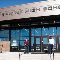 Ivan and Marci stand on the front steps of West Jessamine High School. Both are holding onto the handrails of the steps and smiling.