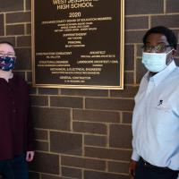 Ivan and Marci stand next to a West Jessamine High School plaque that’s attached to a brick wall inside the school. Both are wearing masks and looking at the camera.