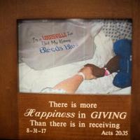 A photo in a frame, showing Ivan and Marci at the hospital. They are holding hands and wearing hospital gowns. Only their arms are visible. Draped over Ivan is a tee shirt that reads “I’m a Louisville fan but my kidney bleeds blue.” The photo frame says “There is more happiness in giving than there is in receiving.”