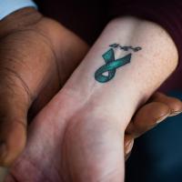 A close-up of the inside of Marci’s wrist, which features a tattoo of a green ribbon and the date 8-31-17. Ivan is holding Marci’s wrist in his hand.