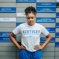 Jaida poses with her hands on her hips, wearing a white University of Kentucky t-shirt, in front of a wall with tiles of names of past University of Kentucky swimmers and divers.
