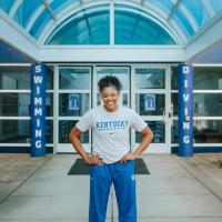 A portrait of Jaida, standing outside the door of the UK Swimming and Diving facility. She is wearing a Kentucky Swimming and Diving tee and blue sweatpants. She has her hands on her hips and is smiling broadly.