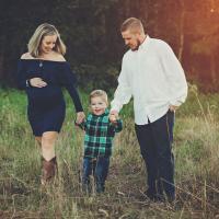 Jennifer Brinegar, her husband Michael Brinegar, a white man with short dark-blonde hair and a beard, and their son Bennett, a young blonde white child, walk hand in hand through a field while on a maternity photo shoot. Michael is wearing a white button-up long sleeve shirt and blue jeans, and Bennett is wearing a blue and green plaid long-sleeve button-up shirt and blue jeans.