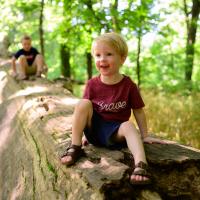 Jeremiah, now a toddler, sits and smiles on a log out in the woods. Another child can be seen playing on the same log behind Jeremiah.