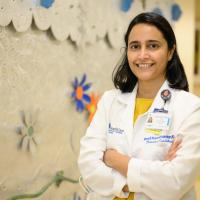Dr. Preeti Ramachandran, a young South Asian woman with medium-length black hair, stands in front of a mural with her arms crossed as she smiles for the camera. She is wearing a white lab coat that has her credentials on it overtop of a yellow shirt, and she is wearing a pair of pearl earrings.