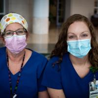 Two female nurses sit next to each other. Both are wearing masks and blue scrubs.