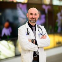 A photo of Dr. Andrew Kolodziej smiling and posing with his arms crossed in front of a mural at the hospital. He is a bald white man with a well-kept dark but graying beard. He is wearing a white lab coat over a black button-up shirt with a gray tie.