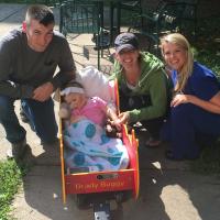 A photo of Max, Carly, and one of Kailey’s caretakers posing outside with Kailey as she sits in a wagon called the “Brady Buggy.”