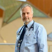 A photo of Dr. Stefan Kiessling posing for the camera in front of a stained glass piece in the hospital. Dr. Kiessling is an older white man with short gray hair and a short gray goatee. He is wearing a long-sleeve light blue button-up with a dark blue tie and a stethoscope around his neck.
