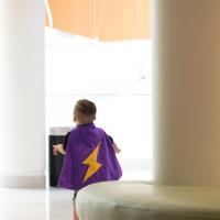 Kase is walking away wearing a purple cape decorated with a large yellow lightning bolt on his back.