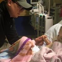 Leorah’s father, a white adult man wearing a black windbreaker, hat, and glasses, consoles her while she sits in her hospital bed and clutches her blanket.