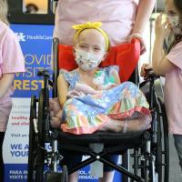 Leorah sits in her wheelchair, legs crossed and visibly smiling from behind her medical mask, with two young girls by her side. Both of these young girls are white, wearing pink “Lenorah Strong” t-shirts and jeans and both have medium-length hair, one blonde and one brunette.