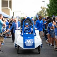A little boy, riding in a futuristic-looking cart, leads Coach Stoops, Courtney Love, and the football team through a crowd of fans.