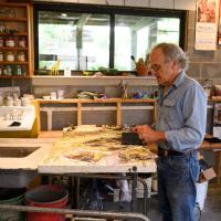 We see Lynn standing at a worktable in his studio in front of an in-progress painting. The painting has several reference photos on it.