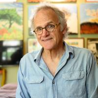 Lynn, smiling, stands in front of a wall of his paintings in his studio.