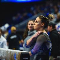 Mac, wearing a sparkly blue, black and white leotard, looks up at something outside of the frame. Her brown hair is up in a bun and has a blue ribbon in it. We can see a blurry crowd behind her.