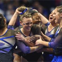 Mac celebrates with a group of very excited gymnasts, all wearing matching blue and black leotards. They are all smiling, jumping, and hugging Mac.