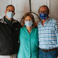 Mae standing closely between her sons. On her left is Kenneth, a middle-aged white man wearing a black jacket, glasses and a facemask. On her right is Scotty, in a white and blue checkered shirt, black framed glasses and a facemask.